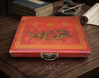 Puede incluir: Una caja de almacenamiento rectangular roja con una ilustración dorada de un dragón y un fénix en la tapa. La tapa muestra tres caracteres chinos dorados y un borde decorativo. Un cierre metálico está centrado en el borde frontal.