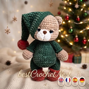 Op de afbeelding: Een gehaakte teddybeer met lichtbruine vacht, een witte snuit en een donkerbruine neus. De beer draagt een donkergroene outfit met capuchon, een rode pompon op de capuchon en bijpassende rode schoenen. De tekst "bestCrochetNest" is onderaan zichtbaar.