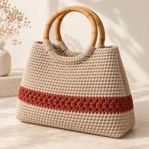 May include: A beige crocheted handbag featuring a horizontal band of dark orange yarn in a braided pattern. The bag has two round, light brown bamboo handles attached to the top. The body of the bag is constructed with a dense, textured stitch.
