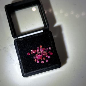 May include: A collection of small, round, faceted pink and red gemstones arranged on a black foam insert inside a square black plastic display box with a clear hinged lid. The stones exhibit varying shades of magenta, crimson, and light pink.
