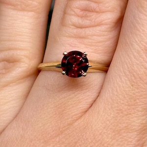 May include: A gold ring featuring a round, deep red faceted gemstone set in a four-prong mounting. The band is smooth and polished, showcasing a classic design with a vibrant, dark red stone as the central focal point of the jewellery piece.