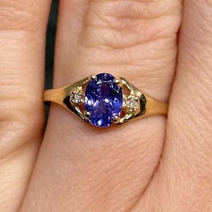 May include: A gold ring featuring a central oval-cut purple-blue gemstone held by four prongs, flanked by two small clear round stones on either side of the setting. The band is smooth and polished, showcasing a classic design with a vibrant centre stone.