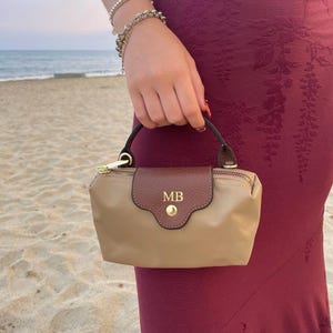 Puede incluir: Un bolso pequeño de color beige con una solapa de cuero texturizado marrón oscuro que presenta las iniciales MB en dorado y un botón de presión de metal dorado. El bolso tiene un asa marrón oscuro, cremallera dorada y cuerpo de nailon.