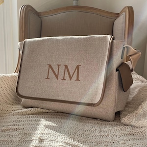 May include: A beige textured messenger bag with a flap closure featuring the letters NM in a dark brown serif font. The bag has brown piping along the edges and a side pocket with a brown accent panel, constructed from a woven fabric material.