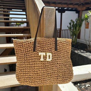 May include: A tan woven straw tote bag featuring two dark brown leather shoulder straps and two white capital letters TD with gold trim centered on the front. The bag has a rectangular shape with a textured surface and a zipper closure at the top.