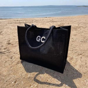May include: A black rectangular tote bag made of a textured woven material. The bag features two thick, rounded black rope handles and the white embroidered letters GC centered on the front panel. The design is structured with a flat base.