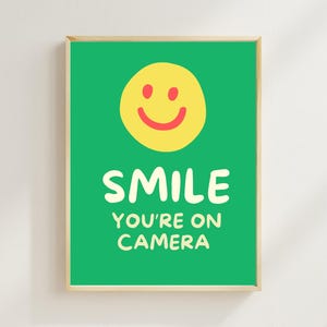 May include: A framed print with a green background and a yellow smiley face. The text "SMILE YOU'RE ON CAMERA" is written in white. The frame is a light wood color.