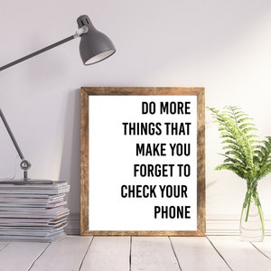 以下が含まれることがあります： 白い背景に黒い文字で「もっと携帯電話をチェックすることを忘れさせるようなことをしよう」と書かれた額装されたプリント。プリントは木製の額縁に入っていて、白い表面に置かれており、左側にランプと雑誌の束、右側に鉢植えの植物がある。