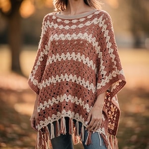 Puede incluir: Un poncho de ganchillo con rayas horizontales en tonos alternos de terracota y crema. La prenda tiene un escote ancho, un patrón de tejido abierto y un dobladillo acabado con largos flecos colgantes en colores a juego.