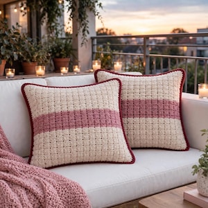 Puede incluir: Dos cojines cuadrados con un patrón de ganchillo texturizado en color crema y una franja horizontal ancha en rosa empolvado. Cada cojín tiene un borde de color granate oscuro a lo largo de los lados, creando un marco para el diseño central.