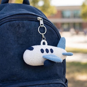 Puede incluir: Un llavero de avión tejido a ganchillo con cuerpo blanco, alas y cola de color azul claro, y detalles en azul oscuro en la ventana de la cabina, las ventanas laterales y el motor. El peluche está unido a una anilla de metal plateado en una mochila azul marino.