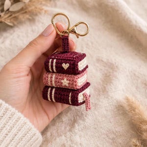 Puede incluir: Un llavero de ganchillo con una pila de tres libros en miniatura en tonos burdeos oscuro y rosa claro. Los libros tienen detalles cosidos en blanco, un pequeño corazón en el libro superior, una estrella en el central y un marcapáginas.