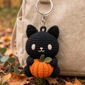 Puede incluir: Un llavero de gato tejido a ganchillo en color negro con cabeza redonda, ojos y boca bordados en blanco, que sostiene una pequeña calabaza tejida en color naranja con un tallo y una hoja verdes. La figura está unida a una argolla metálica.