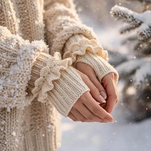 Puede incluir: Un suéter de punto trenzado color crema con mangas largas, puños acanalados y bordes con volantes decorativos. El tejido de punto grueso y texturizado está cubierto por una ligera capa de copos de nieve que resalta el patrón.