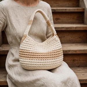 Puede incluir: Un bolso de ganchillo en forma de media luna con un patrón de puntos texturizados en rayas horizontales alternas de color crema y beige claro. El bolso incluye un asa integrada que combina con el esquema de color y el patrón del cuerpo.