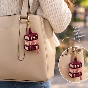 May include: A crochet keychain featuring a stack of three miniature books in shades of burgundy and light pink, attached to a gold-tone metal clasp. The books have embroidered details including a heart and a star, with the text Small details make a big difference.