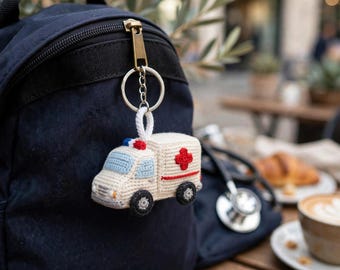 May include: A cream-colored crocheted ambulance keychain featuring light blue windows, a red cross emblem on the side, and a small red and blue light on the roof. The soft, yarn-based vehicle is attached to a silver metal split ring keychain.