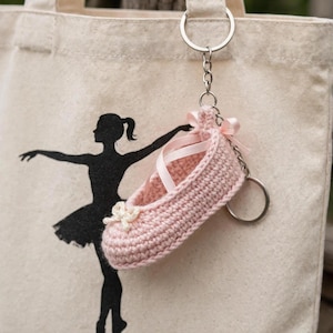 May include: A light pink crocheted ballet slipper keyring featuring a small white bow at the toe and thin pink ribbon laces. The keyring is attached to a silver metal ring and chain, hanging against a beige canvas tote bag with a black dancer silhouette.