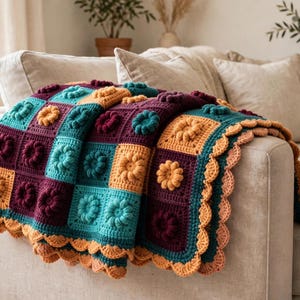 Puede incluir: Una manta de ganchillo hecha a mano con cuadrados en tonos ciruela, azul cerceta y amarillo mostaza. Cada cuadrado presenta un motivo floral en relieve. La manta está rematada con un borde festoneado en color melocotón.
