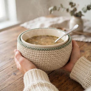 Puede incluir: Un cuenco de cerámica lleno de sopa humeante, encajado en una funda de ganchillo beige gruesa con un borde trenzado de color verde salvia. Una cuchara de metal plateado descansa dentro del cuenco, sostenido por manos con un suéter de punto crema.