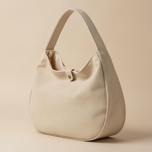 Puede incluir: Un bolso hobo de lona beige con una sola correa para el hombro y un cierre central de botón a presión. El bolso presenta una silueta curva en forma de media luna con un exterior de tela texturizada y un forro interior liso visible.