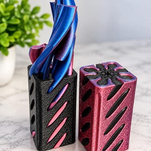 May include: Two rectangular organizers with diagonal slots and a flower-shaped top opening. One organizer is matte black with metallic blue and magenta inserts, while the other is metallic magenta with black inserts. Blue and magenta tools are inside.