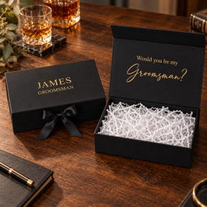May include: Two black rectangular gift boxes with magnetic closures. One box is closed with a black ribbon and features the gold text JAMES GROOMSMAN. The open box contains white crinkle paper and displays the gold text Would you be my Groomsman?