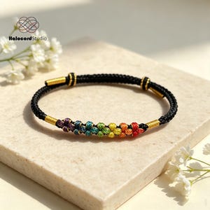 May include: A black cord bracelet featuring a series of small, multi-coloured flower-shaped knots in a rainbow gradient. The bracelet includes metallic gold thread accents at the ends and near the floral centrepiece, with a sliding adjustable closure.