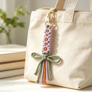 May include: A macrame keychain featuring a vertical row of four lavender flowers with orange centers, attached to a gold-toned metal lobster clasp. The design includes a sage green cord tied into a bow at the base with dangling fringe ends.