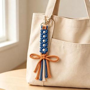 May include: A macrame bag charm featuring a vertical row of blue and white flower-shaped knots attached to a gold-toned metal lobster clasp. The bottom is finished with an orange cord bow and dangling orange, blue, and white cord fringe ends.