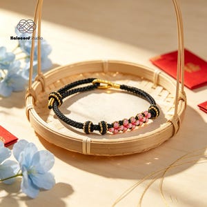 May include: A black cord bracelet featuring a series of pink woven flower-shaped knots and small gold-toned beads. The bracelet includes an adjustable sliding knot mechanism and is branded with the Halocord Studio logo in the upper left corner.