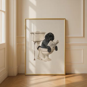 Puede incluir: Una lámina de arte enmarcada que muestra a un perro negro de pelo rizado sentado en un inodoro de porcelana blanca de estilo vintage mientras sostiene y lee un periódico abierto. La obra está en un marco fino dorado con fondo neutro.