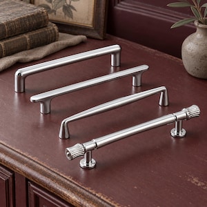 May include: Four silver-toned metal cabinet pulls arranged in a row. The first three pulls feature smooth, cylindrical bars with rounded ends, while the fourth pull has a textured, ribbed design on both ends of the handle.