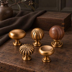 May include: A collection of five cabinet knobs featuring various shapes and finishes. Included are two smooth brass knobs, two fluted brass knobs with radial patterns, and one spherical knob crafted from dark brown wood with a brass base.