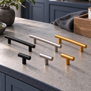 May include: A collection of six cabinet hardware pieces including three long bar pulls and three T-shaped knobs. The hardware is finished in matte black, brushed silver, and brushed gold, featuring a cylindrical design with two mounting posts.