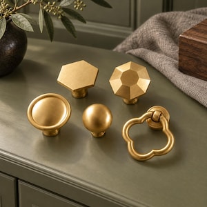 May include: A collection of five brass cabinet knobs and pulls in various shapes, including a hexagon, a faceted octagon, a round disc, a small sphere, and a clover-shaped ring pull, all featuring a smooth, matte metallic finish.