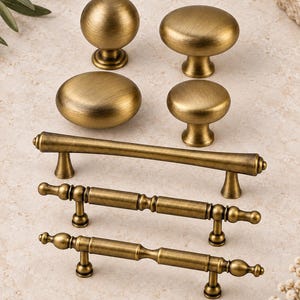 May include: A collection of seven brass cabinet hardware pieces featuring a brushed finish. The set includes four round and oval knobs of varying sizes and three horizontal drawer pulls with distinct cylindrical and tapered design elements.
