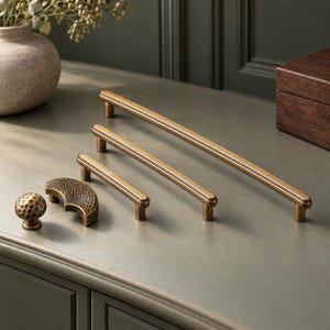 May include: A collection of antique brass hardware including three linear cabinet pulls of varying lengths, a small spherical knob with a hammered texture, and a crescent-shaped pull featuring a matching hammered finish.