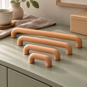 May include: A set of four light-colored wooden cabinet pulls arranged in a cascading size order from longest to shortest. Each handle features a smooth, rounded U-shaped design with a natural wood grain finish, positioned on a flat surface.