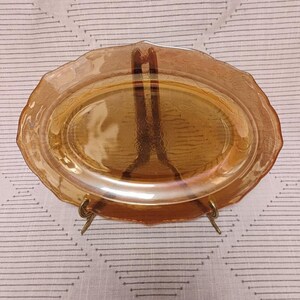Könnte beinhalten: Eine ovale Servierplatte aus bernsteinfarbenem Glas mit gewelltem Rand und einem strukturierten Blumenmuster entlang der Kante. Die Platte steht aufrecht auf einem einfachen Metallständer und zeigt ihre Transparenz und goldene Farbe.
