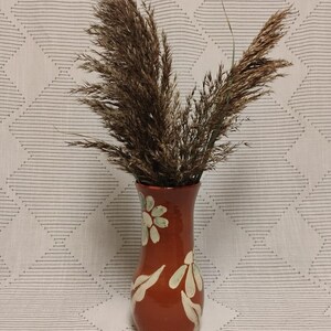 May include: A glossy terracotta-colored ceramic vase with a narrow neck and a wider base, featuring hand-painted cream-colored floral motifs. The vase is filled with a cluster of tall, feathery brown dried grasses that fan out at the top.