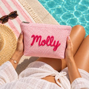 Könnte beinhalten: Eine rechteckige Reißverschlusstasche aus rosa-weißem Seersucker-Stoff. Auf der Vorderseite der Tasche ist der Name Molly in dicken, leuchtend rosa Garnbuchstaben aufgestickt. Die Tasche hat einen silberfarbenen Metallreißverschluss an der Oberseite.