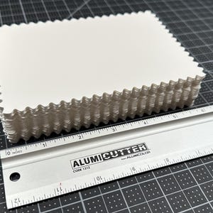 May include: A stack of rectangular white cards with decorative scalloped edges. The cards are positioned next to a silver metal Alumicutter ruler that shows the stack is approximately 3.5 inches wide and 2.5 inches tall in its orientation.