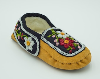 May include: A tan suede moccasin slipper featuring a black suede collar and vamp panel. The black panels are decorated with intricate floral beadwork in white, red, and green. The interior is lined with a cream-coloured fleece material.