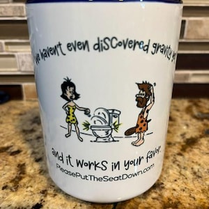 May include: A white ceramic mug with a dark blue rim featuring a cartoon of two cave people near a toilet. The text reads: We haven't even discovered gravity yet and it works in your favor. PleasePutTheSeatDown.com is printed at the bottom.