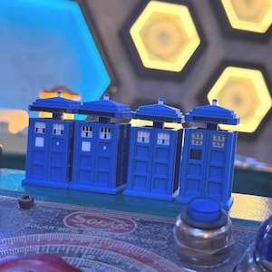 May include: Four small blue police box figurines arranged in a row. Each box features a flat roof, white window panes, and a rectangular door design. The figurines are positioned on a dark control panel with a circular dial and a blue button.