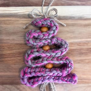 May include: A Christmas tree ornament crafted from a braided, chunky yarn in shades of magenta and gray. The yarn is coiled into a triangular tree shape, accented with three round, natural wooden beads. A jute twine loop and bow are attached to the top.