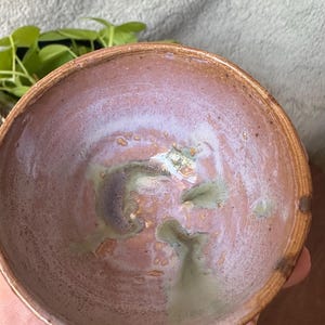 May include: A round ceramic bowl with a thick, textured rim. The interior features a mottled glaze in shades of dusty rose, soft lavender, and muted sage green, with a smooth, glossy finish that pools in the center of the basin.