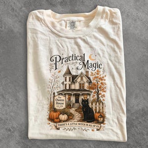 May include: A cream-colored t-shirt featuring a graphic of a Victorian house labeled The Owens House EST. 1998, a black cat, pumpkins, and a lantern. The text reads Practical Magic and There's a little witch in all of us with a crescent moon above.