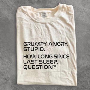 May include: A cream-colored short-sleeve t-shirt with black text printed on the front that reads: GRUMPY. ANGRY. STUPID. HOW LONG SINCE LAST SLEEP, QUESTION? The shirt features a crew neckline and a Comfort Colors brand label at the inner collar.